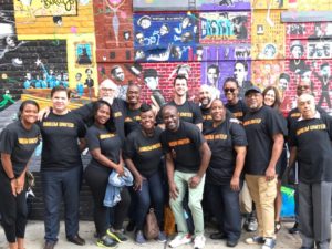 About a dozen people in black t-shirts with the orange Harlem United logo pose for a photograph in front of a colorful mural